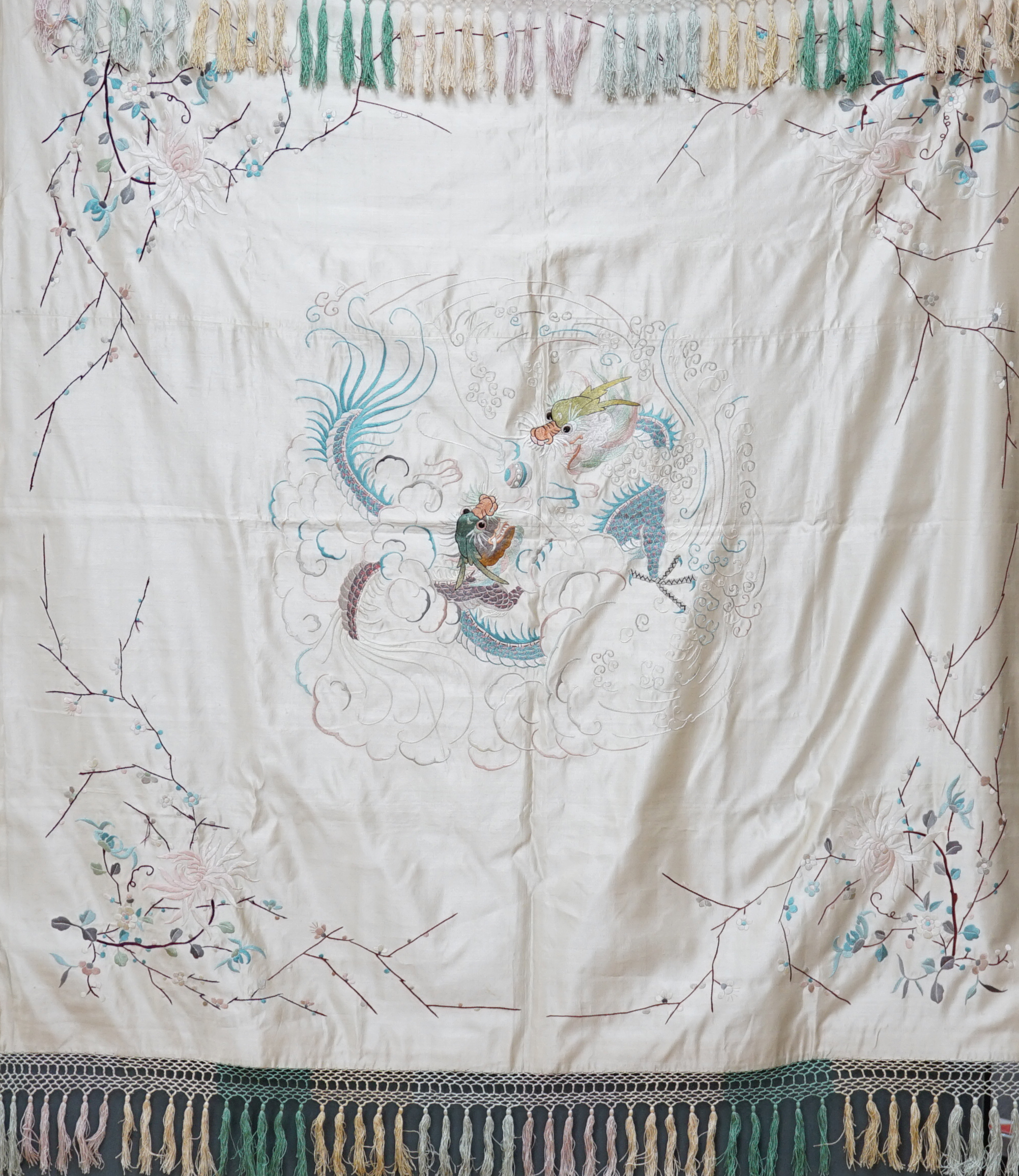 A 1920’s-30’s Chinese oyster silk satin embroidered shawl, now converted to a wall hanging, embroidered in pastel coloured silk threads, the large central motif of two dragons playing with a pearl, the corners of the han